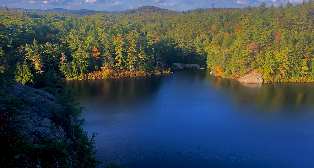 Stonehouse Pond from atop the ledge overlooking the water, a foliage tree line, and mountains in the distance